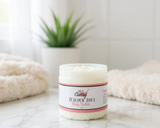 Jar of body butter with a label on a marble surface, with a plant and towel in the background.