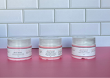 Three jars of body butter on a pink surface with a tiled wall background