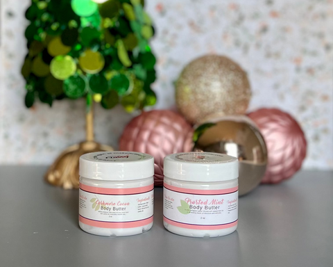 Two jars of body butter on a surface with decorative Christmas tree and ornaments in the background.