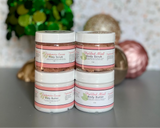 Two jars of body scrub with decorative Christmas tree and ornaments in the background