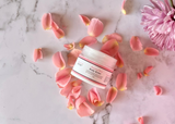 Jar of body butter surrounded by pink flower petals on a marble surface