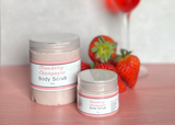 Two jars of strawberry champagne body scrub on a table with strawberries and a glass in the background.