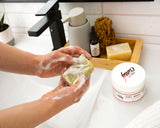 Person washing hands with soap, with a focus on a jar of Blissful Brooklyn Hair Conditioner product on a bathroom counter.